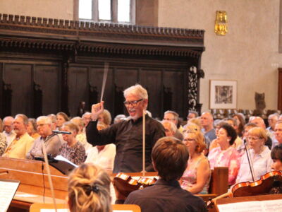 A conductor energetically leads an orchestra in front of a seated audience inside a warmly lit church or hall, with individuals attentively watching the performance.