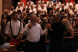A man in a white shirt gives a thumbs-up while an audience applauds enthusiastically in a dimly lit venue, with people in formal attire.