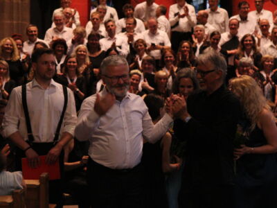 A man in a white shirt gives a thumbs-up while an audience applauds enthusiastically in a dimly lit venue, with people in formal attire.