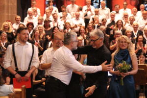 A large choir stands onstage with people in formal attire applauding. Two men are embracing in the center, while a woman holds flowers and smiles.