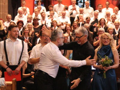 A large choir stands onstage with people in formal attire applauding. Two men are embracing in the center, while a woman holds flowers and smiles.
