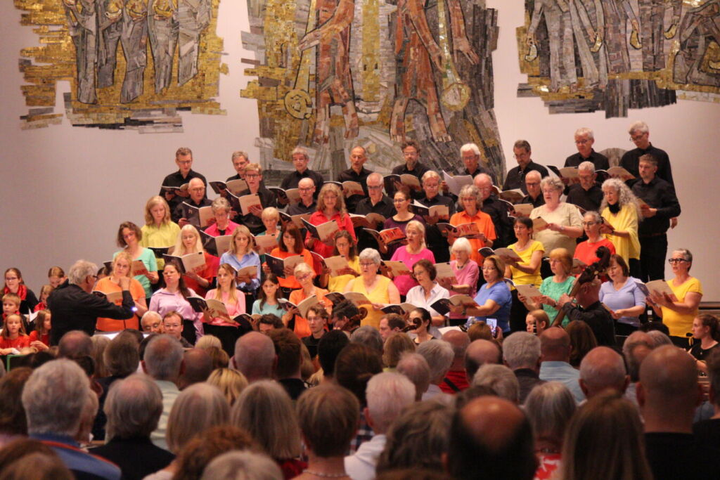 A large choir dressed in colorful shirts performs on stage in front of an audience, with artistic wall decorations in the background.
