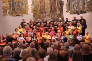 A large choir dressed in colorful shirts performs on stage in front of an audience, with artistic wall decorations in the background.