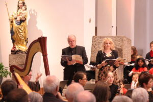 A choir performs in a church, with a man and woman singing from hymnals. A harp is visible in the foreground, and a statue of a saint stands in the background.