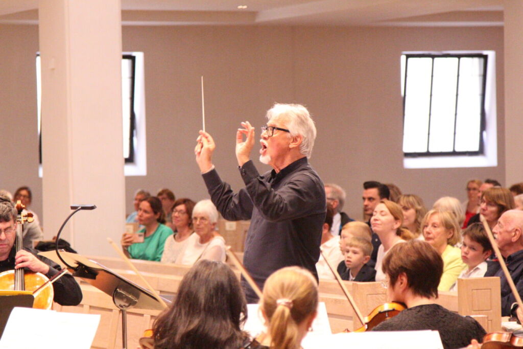 A conductor leading an orchestra in a performance while an audience watches attentively in a well-lit room.