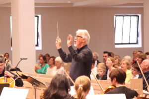 A conductor leading an orchestra in a performance while an audience watches attentively in a well-lit room.