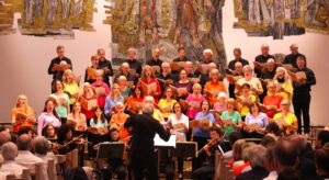 A choir of diverse ages, dressed in colorful shirts, performs in a church setting under the direction of a conductor, with an audience watching attentively.