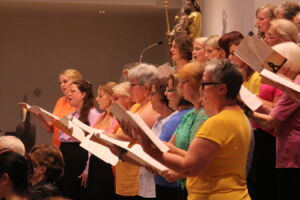A choir of adults in colorful clothing sings in unison, holding sheet music, in a well-lit indoor space.