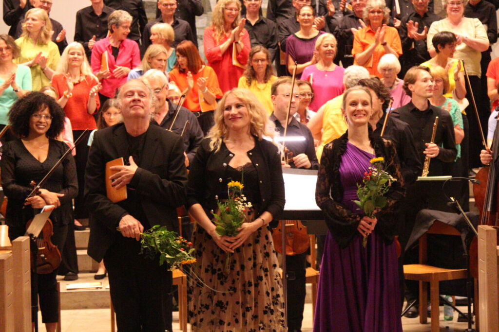 Three people stand at the front of a stage holding bouquets, while a large group of people in colorful attire applaud in the background.