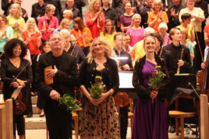 Three people stand at the front of a stage holding bouquets, while a large group of people in colorful attire applaud in the background.