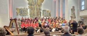 Choir and orchestra performing in a church, with a conductor in front, surrounded by solemn sculptures and an audience.