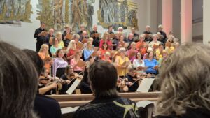 A choir wearing colorful shirts performs in a church setting, accompanied by an orchestra. The audience is seated in the foreground, and the choir stands on risers in front of decorative wall art.