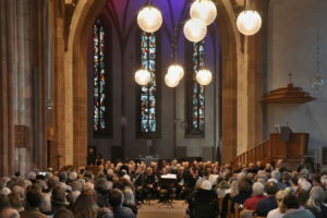 A large audience sits in a church watching a performance, with stained glass windows and hanging lights in the background.