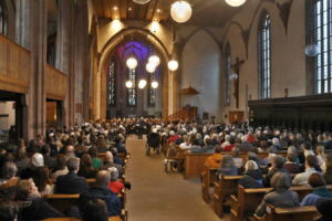 A large congregation sits in a grand church with high ceilings, listening to a speaker at the front. The church is illuminated by large hanging lights and natural light from tall windows.