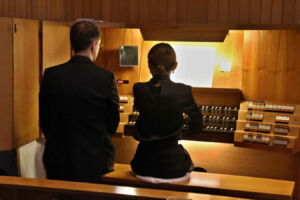 Two people in formal attire sit at a church organ, with one person apparently playing while the other observes, surrounded by wooden panels and softly lit by a lamp.