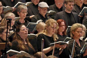 A choir of diverse individuals in black attire, holding songbooks and singing.