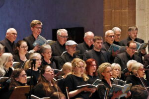 A choir of various-aged singers in black attire performs in a concert setting, holding open songbooks and appearing focused on their music.