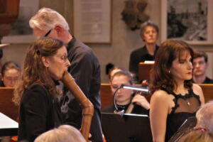Musicians performing in an orchestral setting, with a woman playing a woodwind instrument and others focused on their performance.