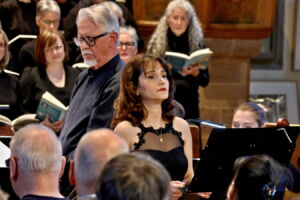 A choir performing in a church, featuring a woman in a black dress and a man with glasses in the foreground, with choir members holding books in the background.