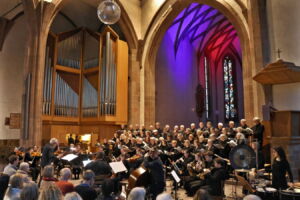 A choral and orchestral performance inside a church, with singers and musicians arranged under colorful arched lighting and a large organ in the background. An audience watches the performance attentively.