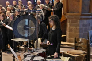 A percussionist stands among drums and a gong, playing in an ensemble. A choir is situated in the background, all in formal attire, singing as part of a performance in a wooden-accented venue.