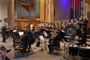 An orchestra performs alongside a large choir in a grand hall with an organ in the background. The conductor stands to the side, guiding the ensemble.