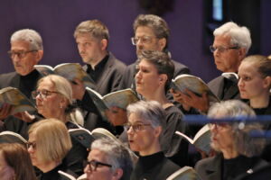 A choir of diverse adults singing passionately from sheet music during a performance.