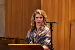 A woman stands at a lectern in an indoor setting, wrapped in a shiny shawl, speaking or singing, with a microphone nearby.
