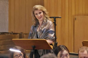 A woman stands at a music stand, wearing a shiny shawl, in what appears to be a church or performance space. A few people are seated in the foreground.