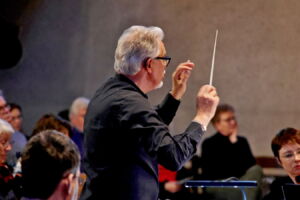 A conductor with gray hair leads an orchestra, holding a baton. The scene captures the side profile of the conductor gesturing with his hands, with orchestra members in the background.