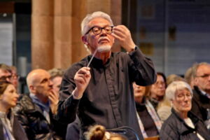 An elderly conductor with gray hair and glasses leads an orchestra indoors, holding a baton. The audience, seated in the background, attentively watches the performance.