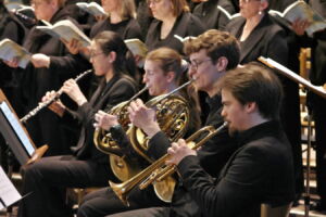 Musicians in formal attire play horn and trumpet alongside a choir reading sheet music during a performance.