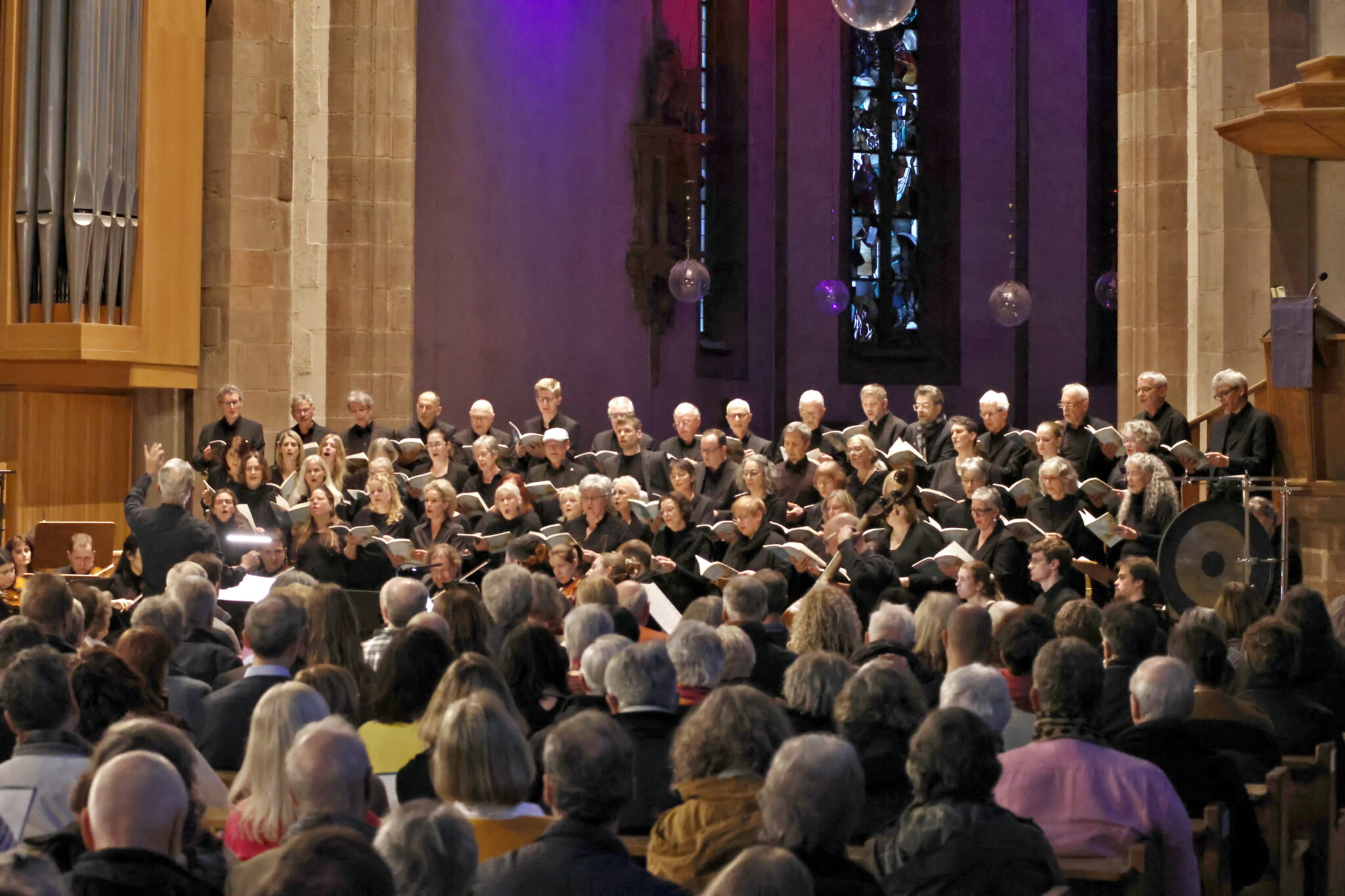 A choir performs in a church with an audience seated in front. The setting has stained glass windows and an organ.
