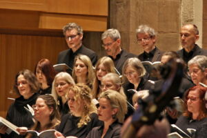 A choir of men and women, dressed in black, is singing during a performance in a concert hall.