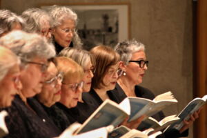 A choir of elderly women singing, each holding an open music book, wearing black outfits, and standing close together in a well-lit room.