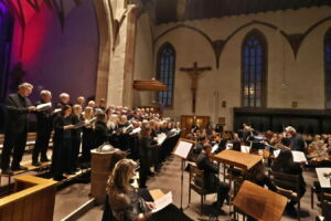 A choir performing with an orchestra in a dimly lit church, featuring stained glass windows and a crucifix in the background.