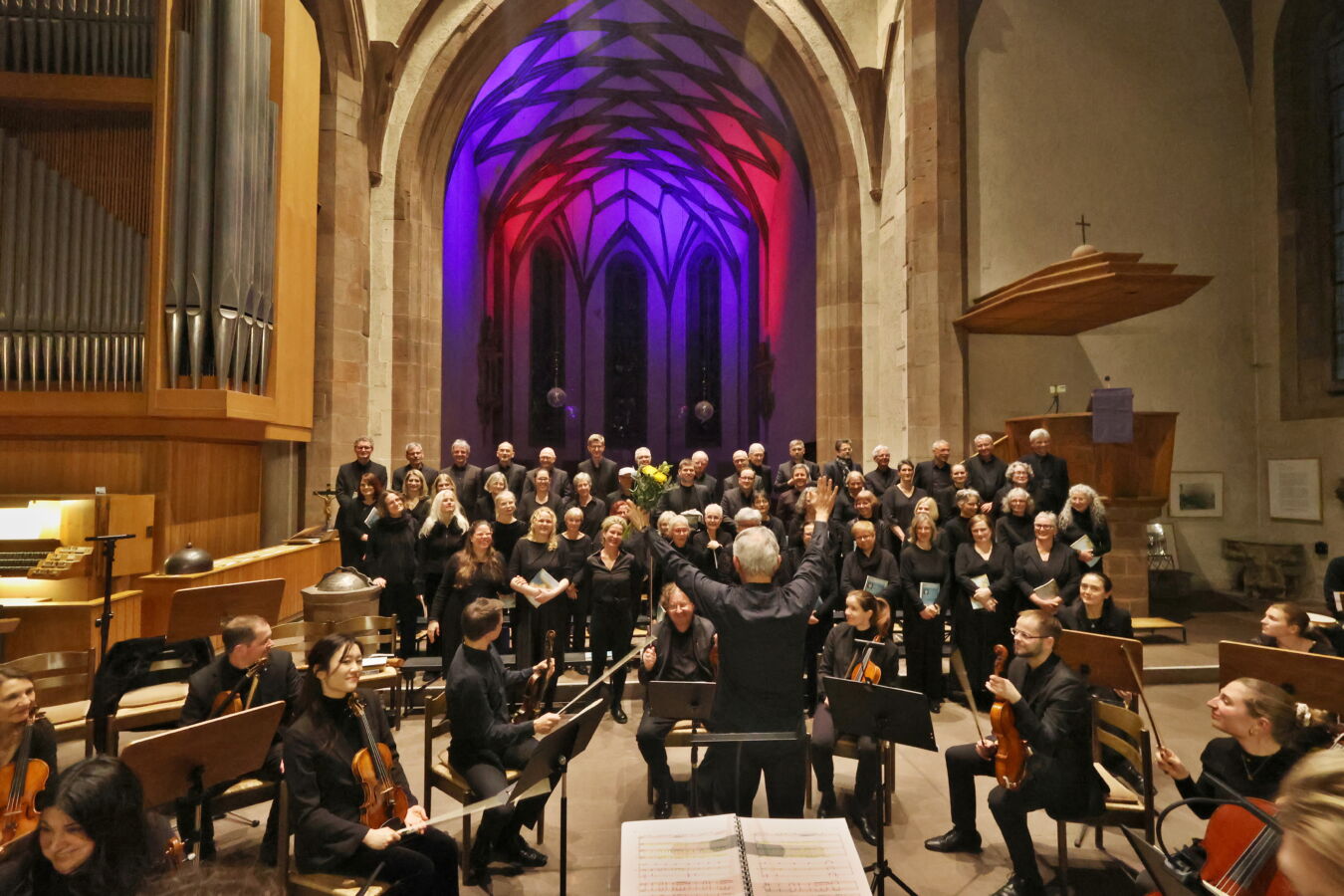 A choir and orchestra perform in a grand cathedral with colorful lighting illuminating the arched ceiling.