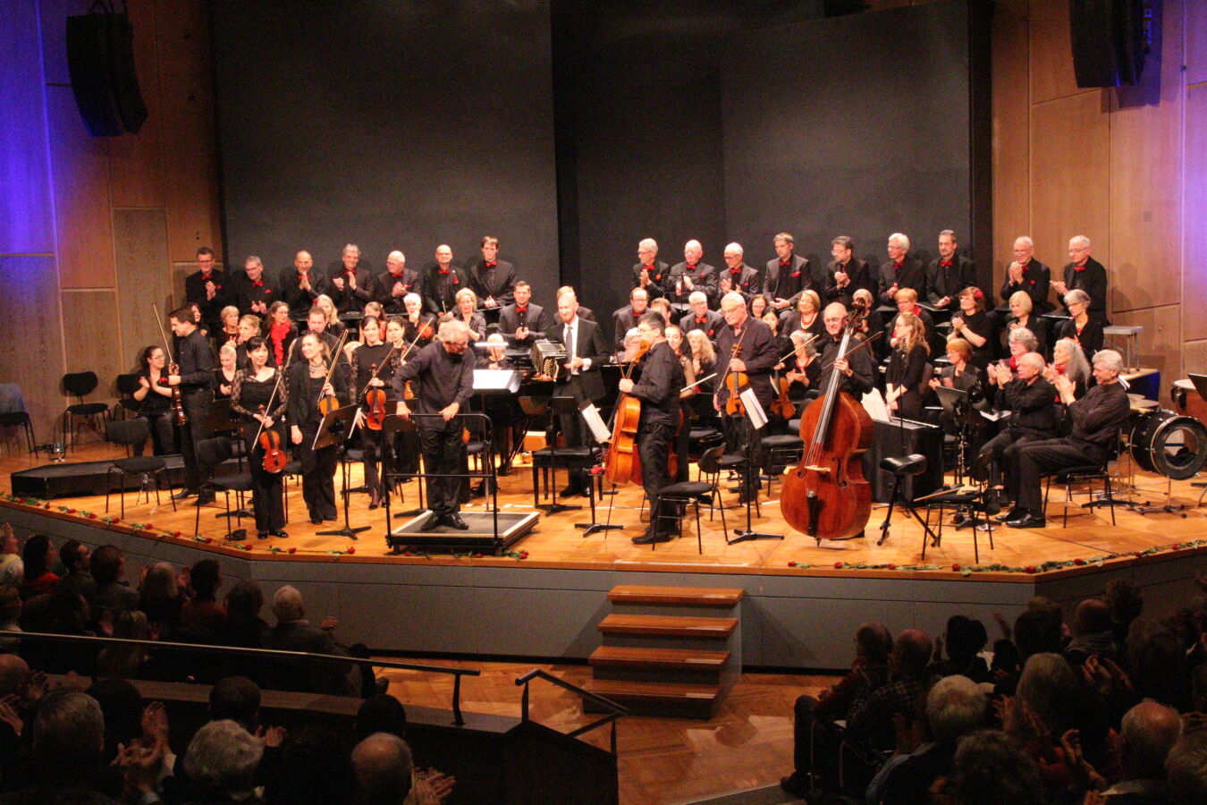 A choir and orchestra perform on a well-lit stage. The musicians, dressed in black, are accompanied by a conductor. An audience is visible in the foreground, attentively watching the performance.
