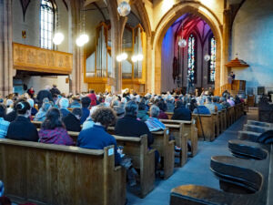 A large congregation sits in wooden pews inside a historic church with high ceilings, stained glass windows, and arched architecture.