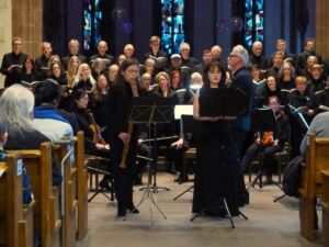 A choir and musicians performing in a church, with stained glass windows in the background and an audience seated in wooden pews.