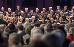 A choir performs on stage in front of a seated audience, with singers holding sheet music and attentively following the conductor.