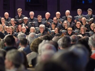 A choir performs on stage in front of a seated audience, with singers holding sheet music and attentively following the conductor.