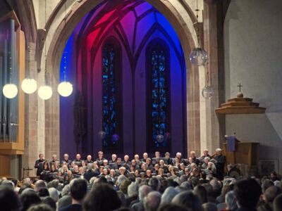A choir performs in a church with a large audience. The arched windows behind the choir are illuminated in red and blue lights, creating a vibrant backdrop.