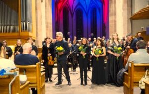 A group of musicians and singers stand on a stage in a church with vibrant backlighting. Some hold yellow flowers, and the audience is seated in wooden pews.