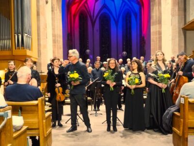 A group of musicians and singers stand on a stage in a church with vibrant backlighting. Some hold yellow flowers, and the audience is seated in wooden pews.