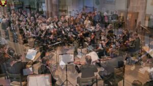 A large orchestra performs in a spacious hall, with musicians playing string instruments and a conductor leading. The audience is seated in the background, listening attentively.
