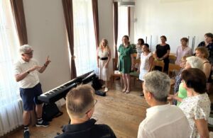 A group of people standing in a room around a piano, with a conductor leading a music session.