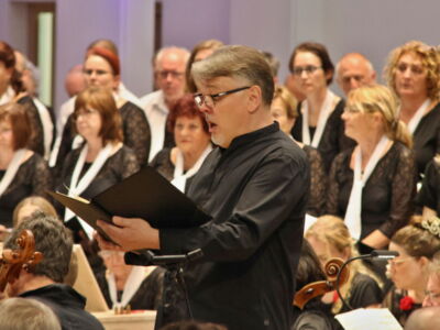 A man in a black outfit sings from a music folder in front of a choir dressed in matching attire during a performance.