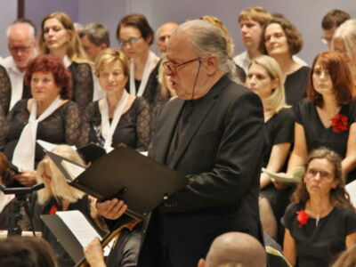 A choir performing, with a conductor in the foreground holding a music score, surrounded by singers dressed in black and white attire.