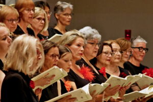 A choir of women in black attire sings passionately from sheet music during a performance.